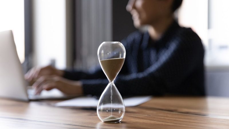 Close up hourglass measuring time, standing on wooden office table, Indian businesswoman working on background, efficiency, deadline and time management concept, busy employee using laptop