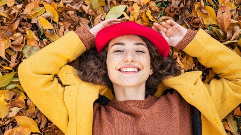 Happy smiling woman lying over yellow leaves and looking at camera. Young beautiful woman lying on dry autumn leaves with a big grin. Portrait of cheerful girl rest on ground in park with hands behind head.