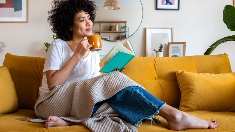 Pensive relaxed African american woman reading a book at home, drinking coffee sitting on the couch. Copy space. Lifestyle concept.
