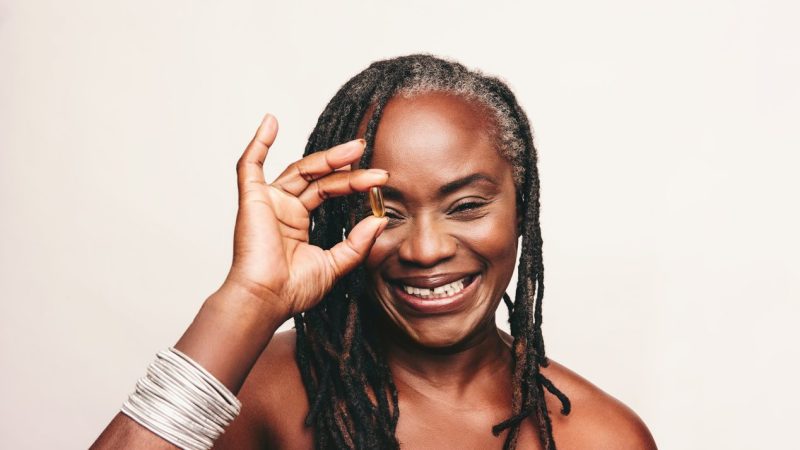 Cheerful mature woman smiling at the camera while holding a capsule of Omega 3 fatty acids. Happy woman with dreadlocks using nutritional supplements as part of a healthy self-care routine.