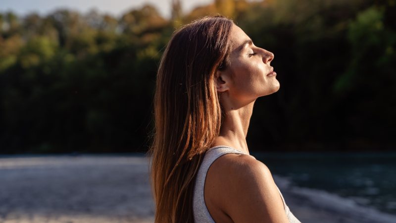 Young woman with long hair enjoying sun with closed eyes getting natural vitamin D outdoors. Peace of mind. Mindfulness, mental health,  spirituality, well-being, unwind yourself