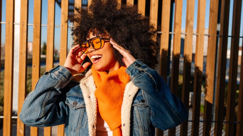 Stylish young woman with afro hair, wearing orange scarf and denim jacket, smiling and adjusting sunglasses against wooden background