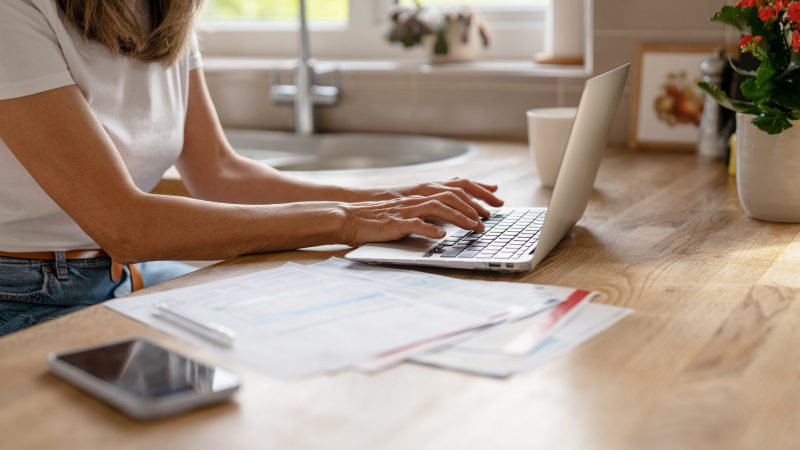 Busy woman focuses on work at her home office, typing on a laptop with paperwork and a smartphone nearby. Bright natural light fills the cozy kitchen space.