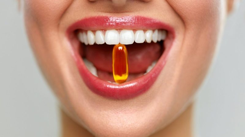 Vitamins And Food Supplements. Close Up Of Beautiful Woman Opened Mouth Holding Fish Oil Pill In White Teeth. Smiling Girl Holding Capsule With Omega-3 Between Teeth. Healthy Diet Nutrition Concept