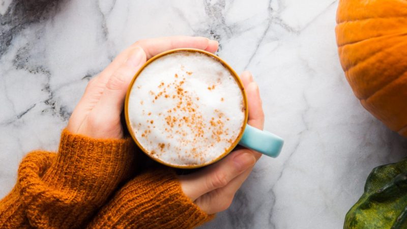 Autumn moody background with mug of latte coffee and pumpkins on marble table. Flat lay in fall colors. Female hands in cozy sweater
